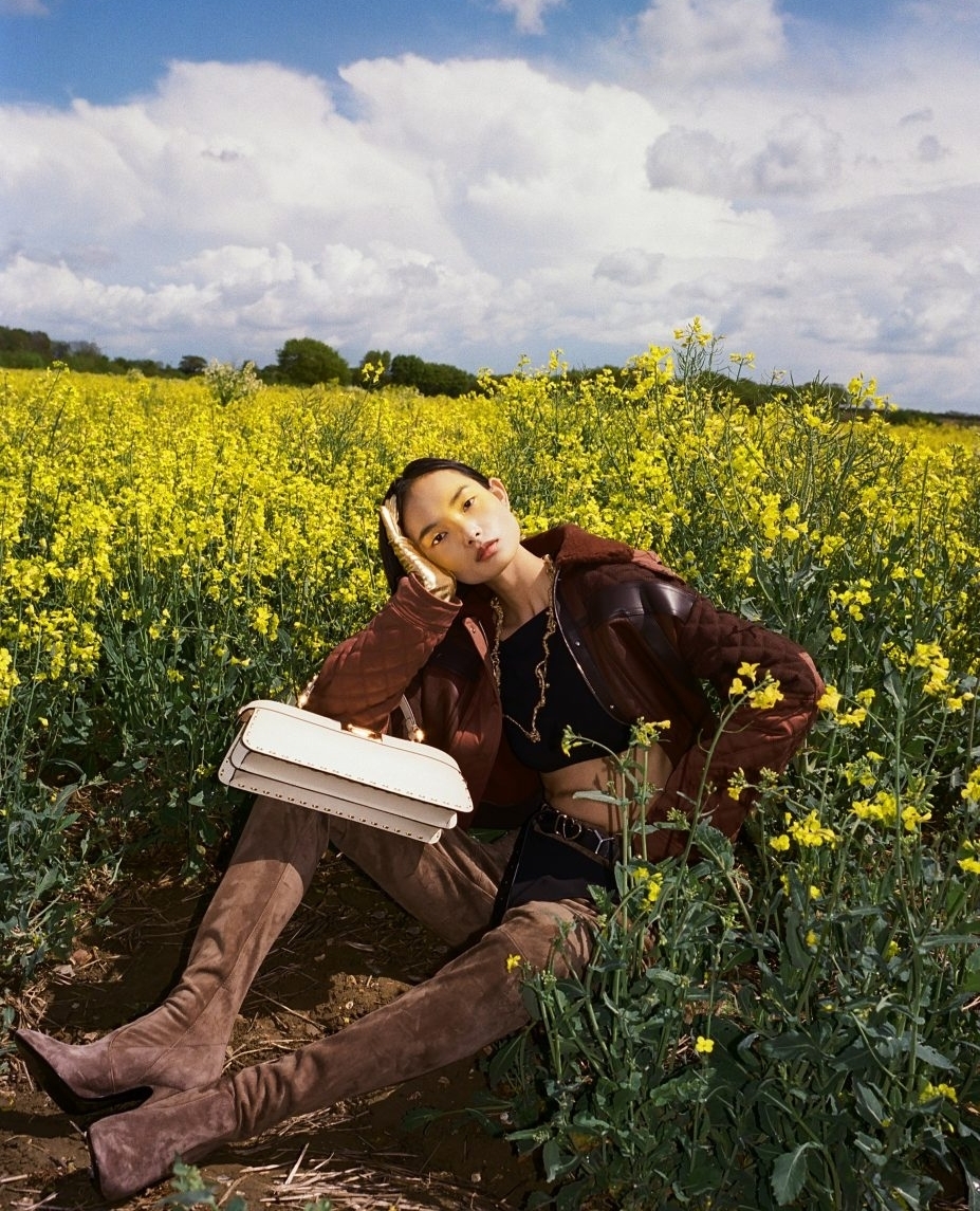 Woman poses in field of yellow flowers wearing a brown leather jacket, thigh-high suede boots, and holding a cream-colored handbag.