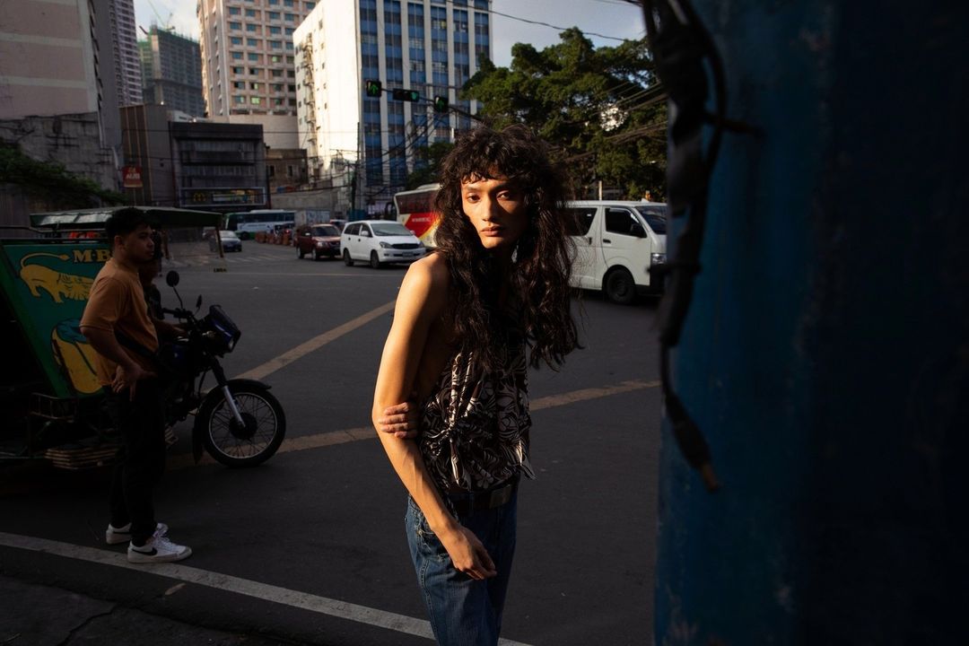 Adult female fashion model with long hair poses on urban street with motorcycle and city buildings during daylight for fashion editorial.