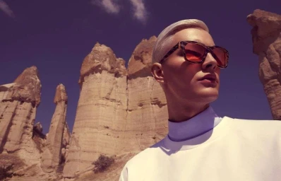 Model wearing patterned sunglasses and white top against tall rock formations under a twilight sky