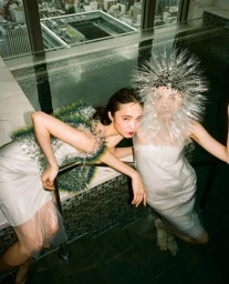 Two women in avant-garde white dresses leaning over an indoor pool with a city view reflected behind them