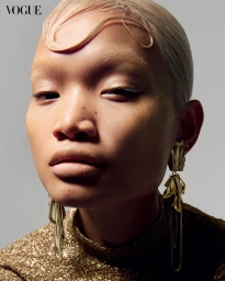 Close-up portrait of a model with sculpted blonde hair, gold metallic top, and ornate gold earrings against a plain grey background