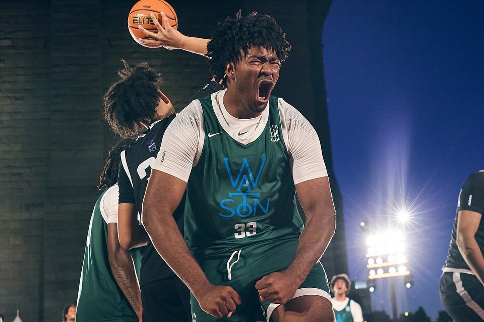 A young, Black basketball player in a green and white jersey shouts in celebration during an outdoor game at dusk, with other players nearby.