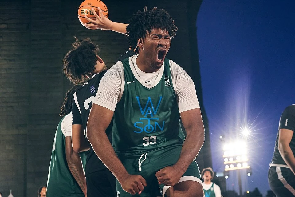 A young, Black basketball player in a green and white jersey shouts in celebration during an outdoor game at dusk, with other players nearby.