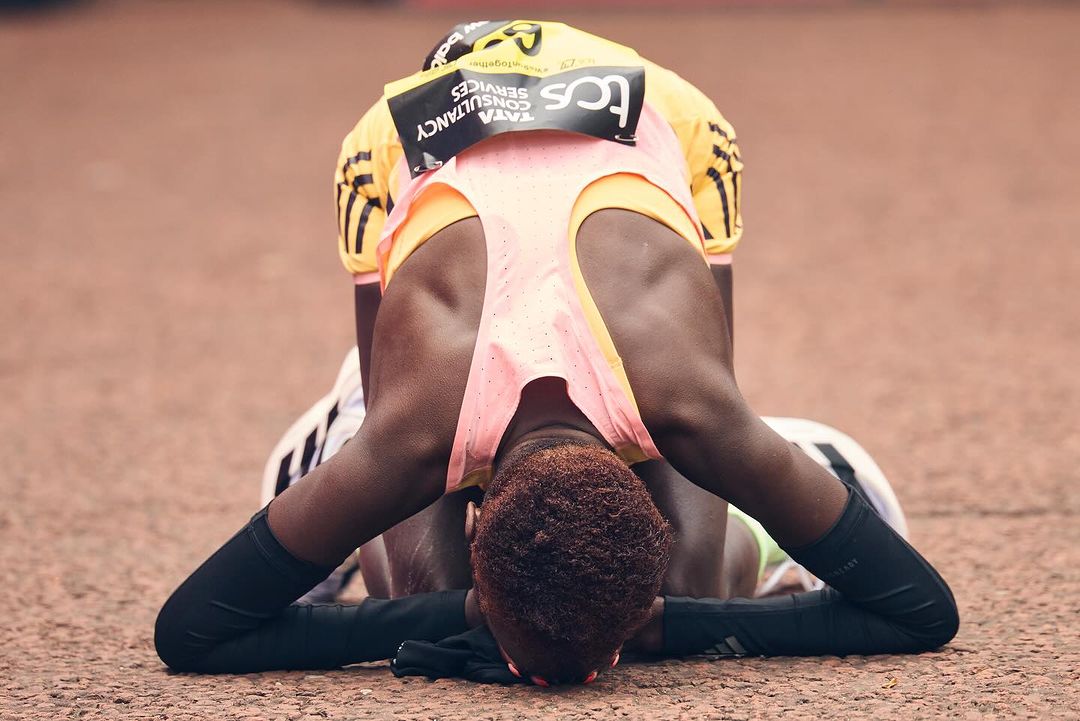 A woman kneels on a red running track in athletic wear, head down, in apparent exhaustion, after a race, with logos visible on her top.
