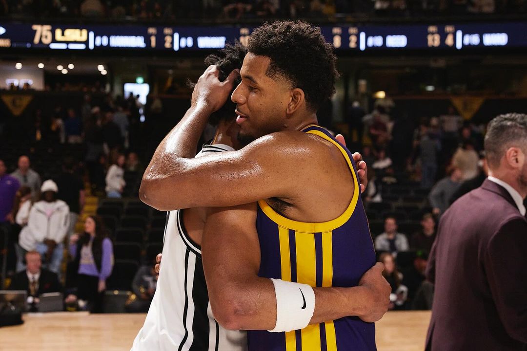 Two basketball players embracing on the court after a game, wearing team uniforms in an indoor stadium setting with crowd.
