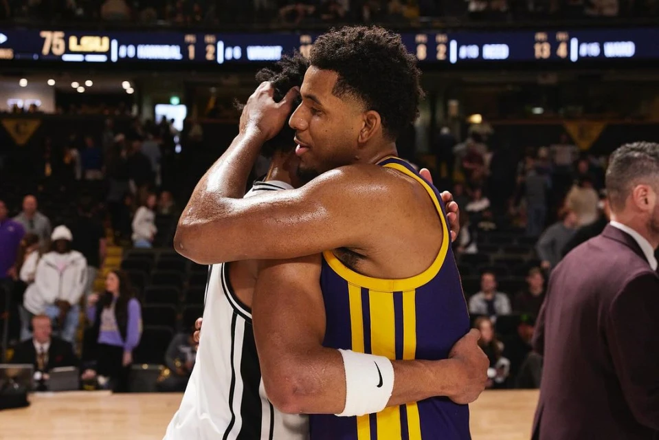 Two basketball players embracing on the court after a game, wearing team uniforms in an indoor stadium setting with crowd.
