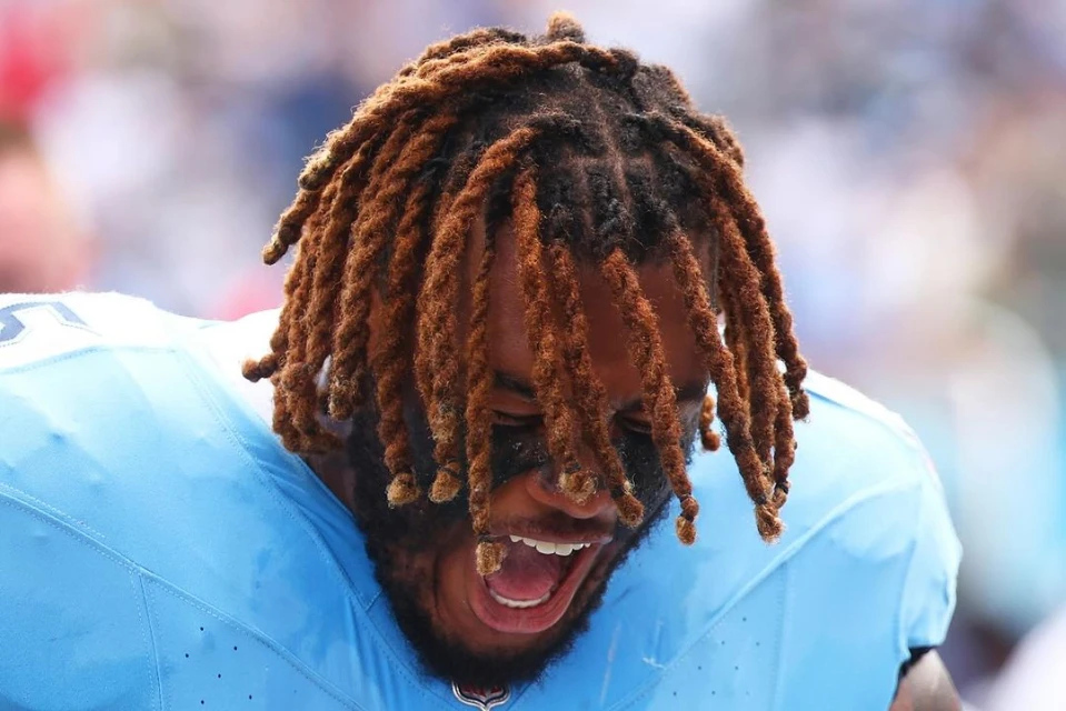 Close-up of an adult male athlete with dreadlocks and black eye paint, wearing a blue sports jersey with an open mouth.