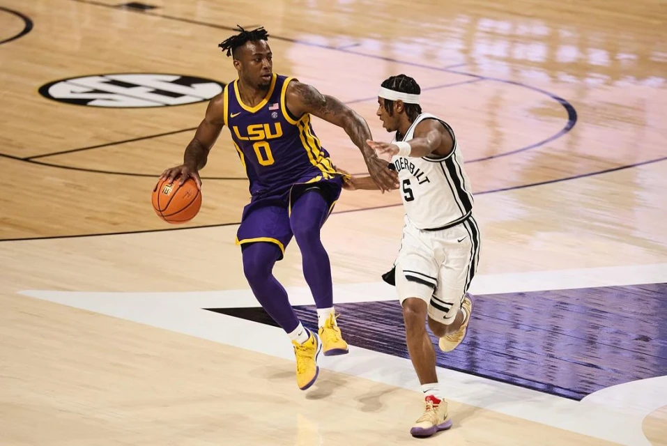 Basketball game action shot featuring two players in uniforms dribbling the ball on a wooden court with stadium seating backdrop.