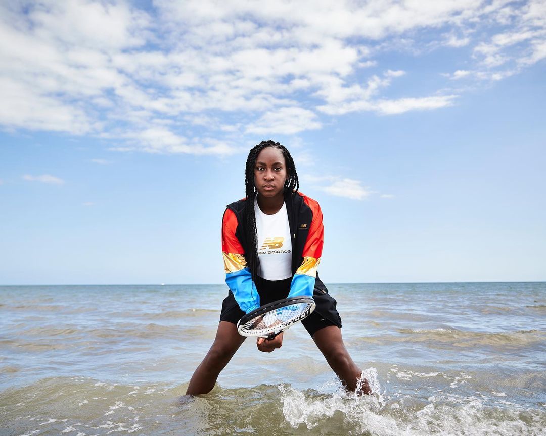 Adult female posing in ocean with racket, wearing athletic clothing and New Balance apparel on a partly cloudy day at the beach.