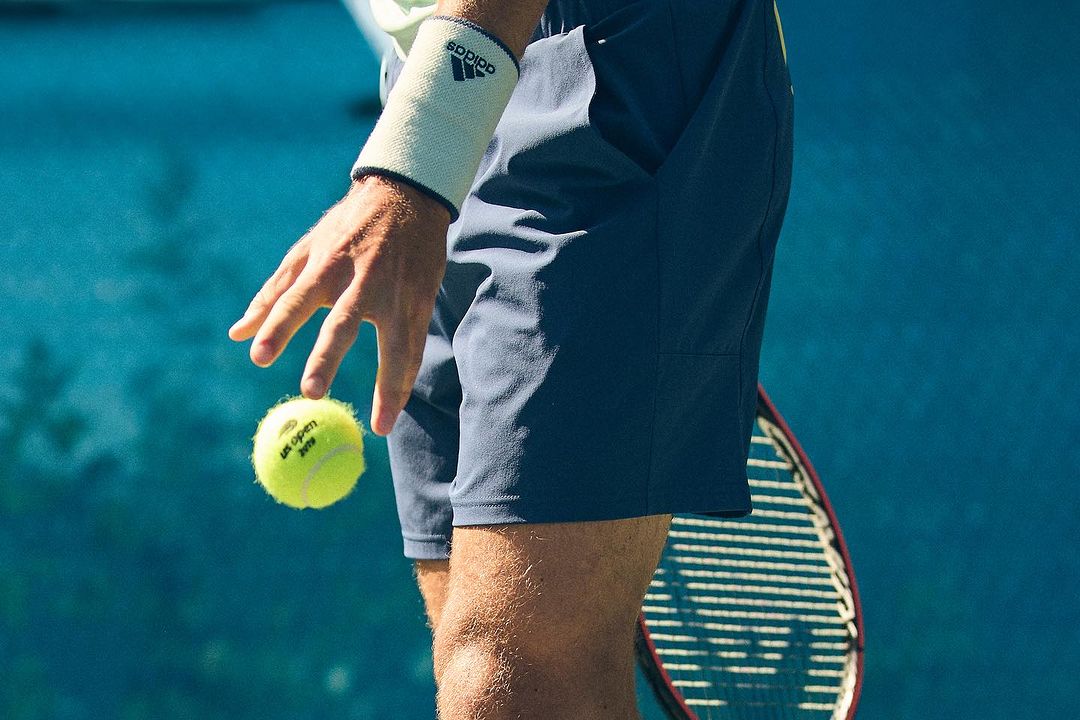Man holding a tennis ball on a blue court, wearing an Adidas wristband and shorts, with a tennis racket visible in the background.