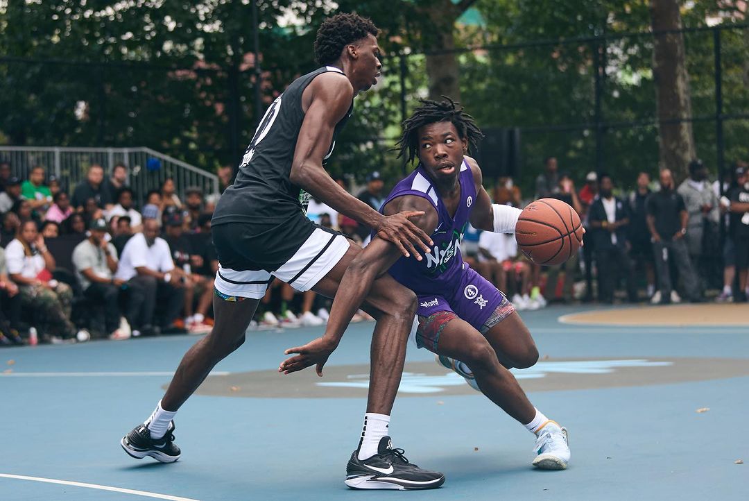 Two young men face off in an intense basketball game on an outdoor court surrounded by spectators, showcasing athletic prowess and competitive spirit.