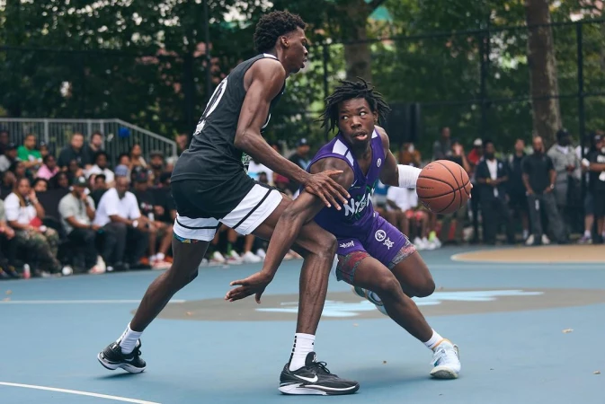 Two young men face off in an intense basketball game on an outdoor court surrounded by spectators, showcasing athletic prowess and competitive spirit.