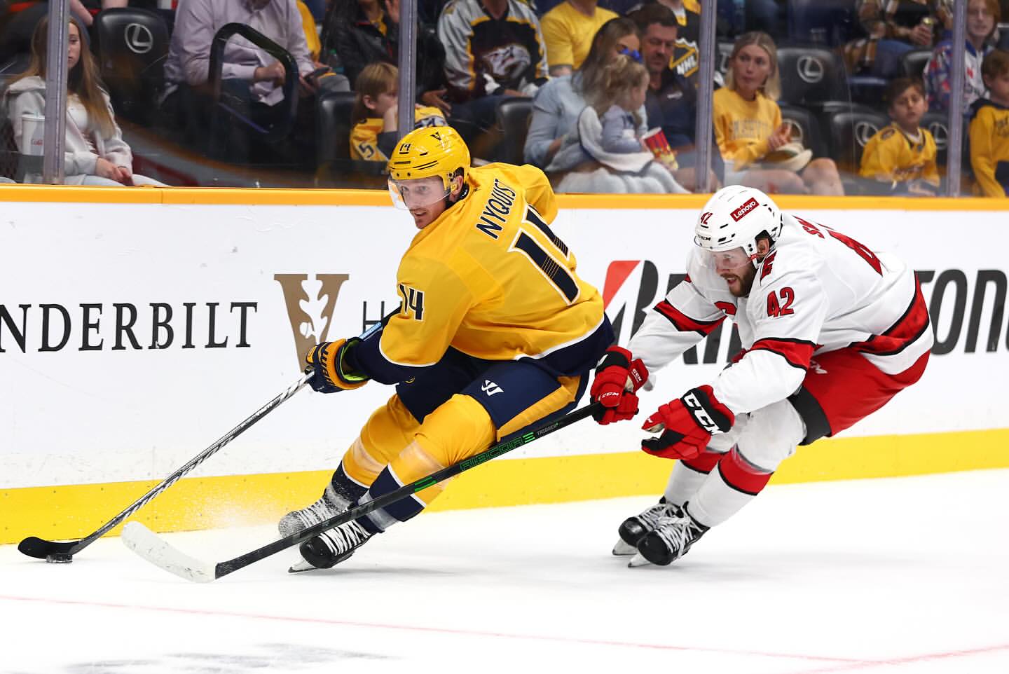 Two hockey players in action on the ice. One in yellow and navy skates towards the camera and another in red and white follows.
