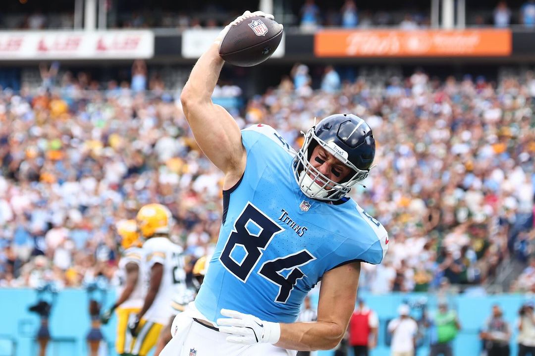 American football player in blue and white uniform raises the ball during a play on a sunny day in outdoor stadium.