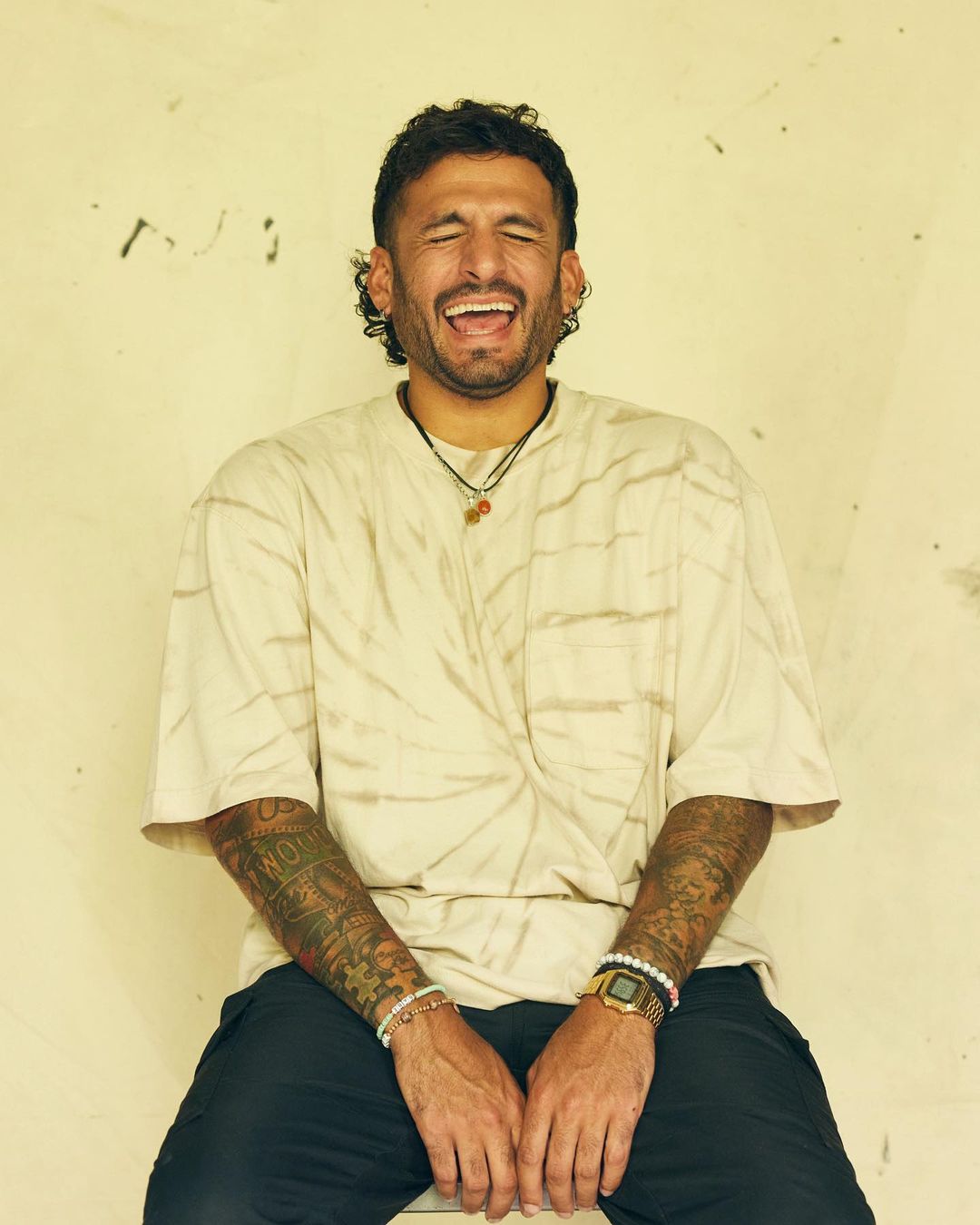 A joyful adult male laughing while sitting indoors, wearing casual clothing and jewelry, with a textured wall as a neutral background.