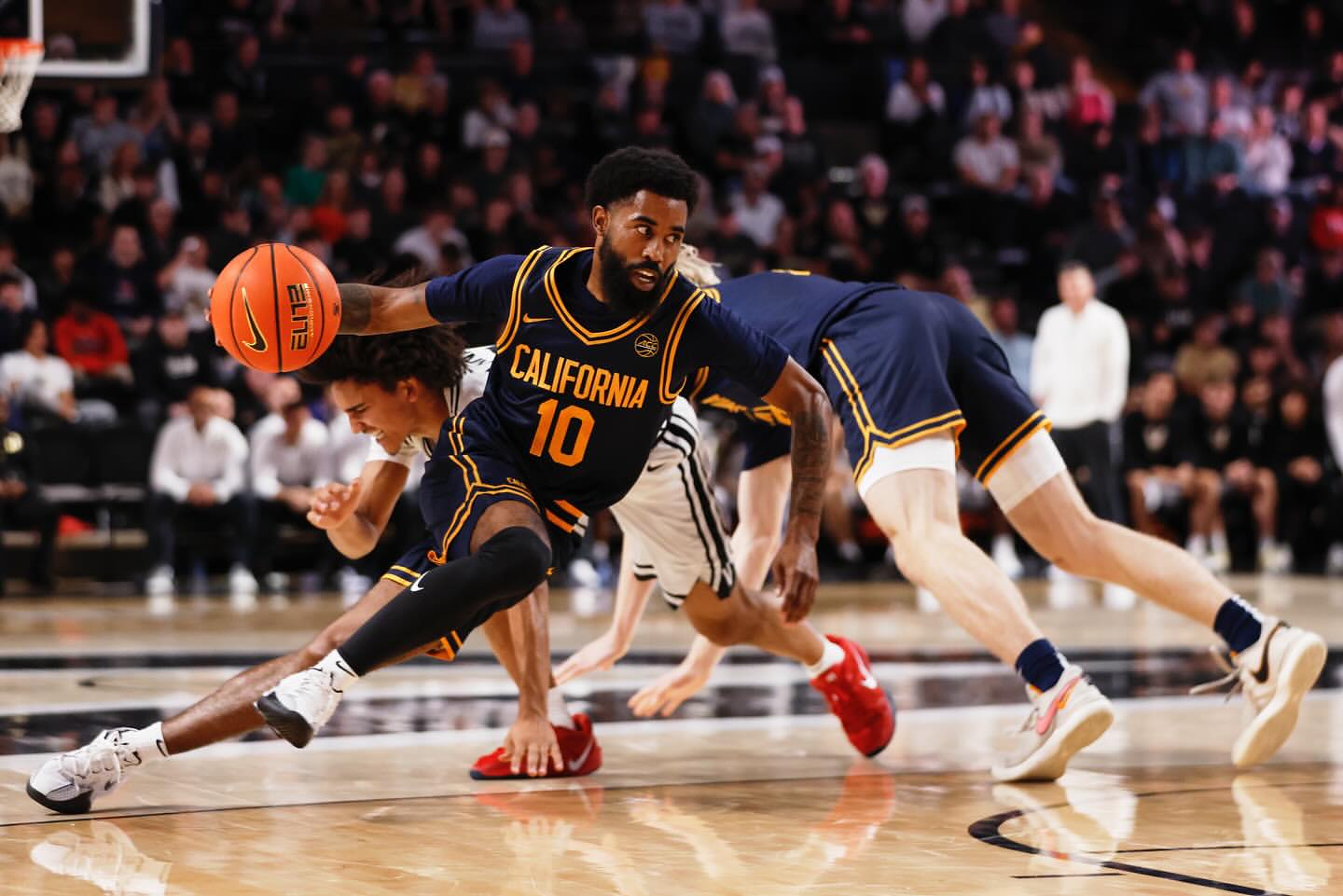 Basketball player with the ball dribbling on the basketball court in a navy and gold uniform; a crowd watches in the background.