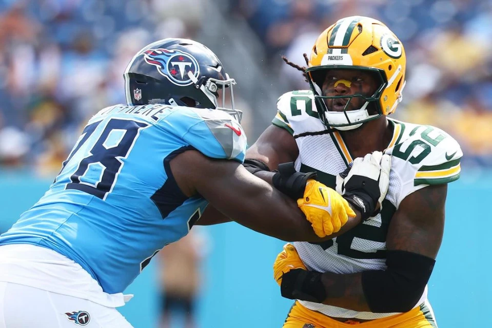 Two American football players in action during a game. One in blue Titans jersey, the other in green Packers uniform, competing closely.