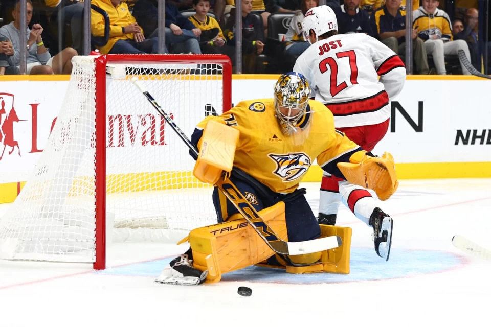 Hockey goalie in yellow prepares to block a shot with another player behind on an ice rink in a brightly lit stadium.