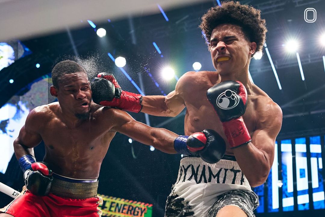 Two shirtless male boxers exchange blows in a boxing ring under bright lights, showcasing intense action in a dynamic sports setting.