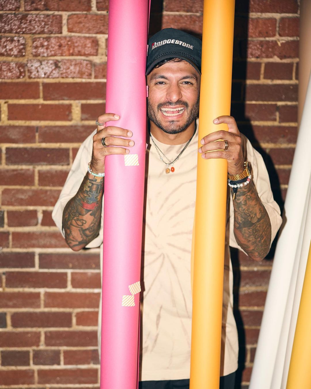 Smiling man holding pink and orange rolls of paper against brick wall, wearing a baseball cap, t-shirt, jewelry, and tattoos.
