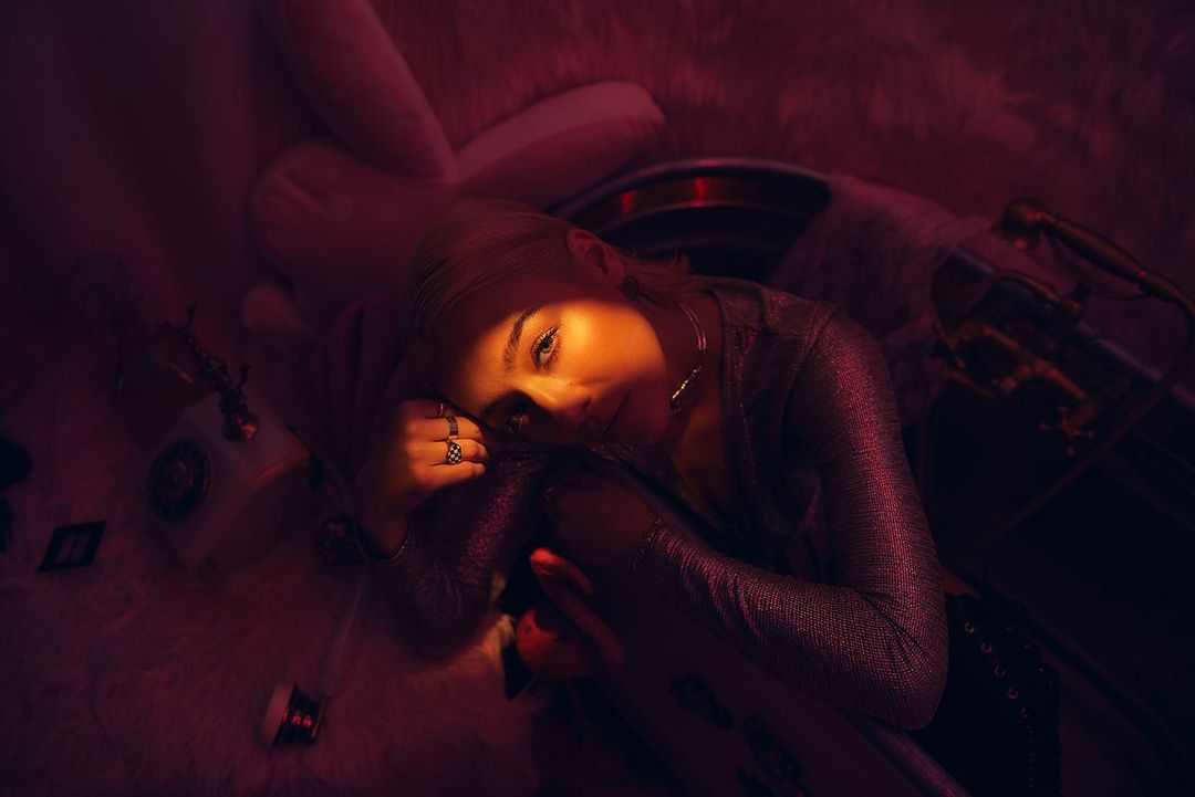 A woman in a long-sleeved shirt rests her head on her knee in a dark, indoor setting illuminated by moody red and orange lighting.