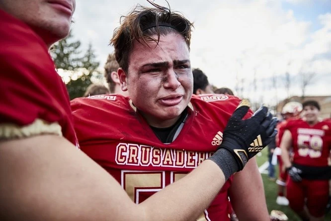 Emotional moment captured of a young male football player in a red uniform after a game, with a teammate's comforting hand on his shoulder.