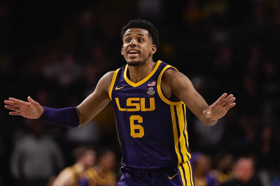 A basketball player in a purple and yellow LSU jersey stands with arms outstretched, looking up during a basketball game.