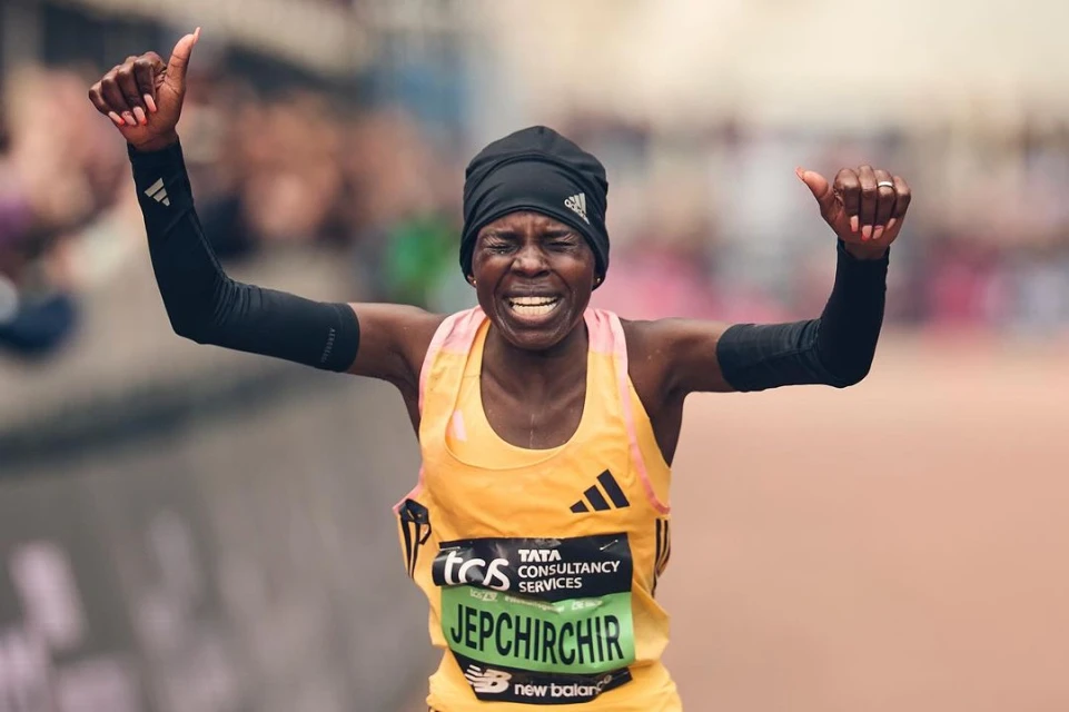 Female marathon runner celebrates victory with raised arms, wearing athletic gear and beanie, cheered on by a blurred background crowd.