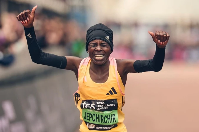 Female marathon runner celebrates victory with raised arms, wearing athletic gear and beanie, cheered on by a blurred background crowd.