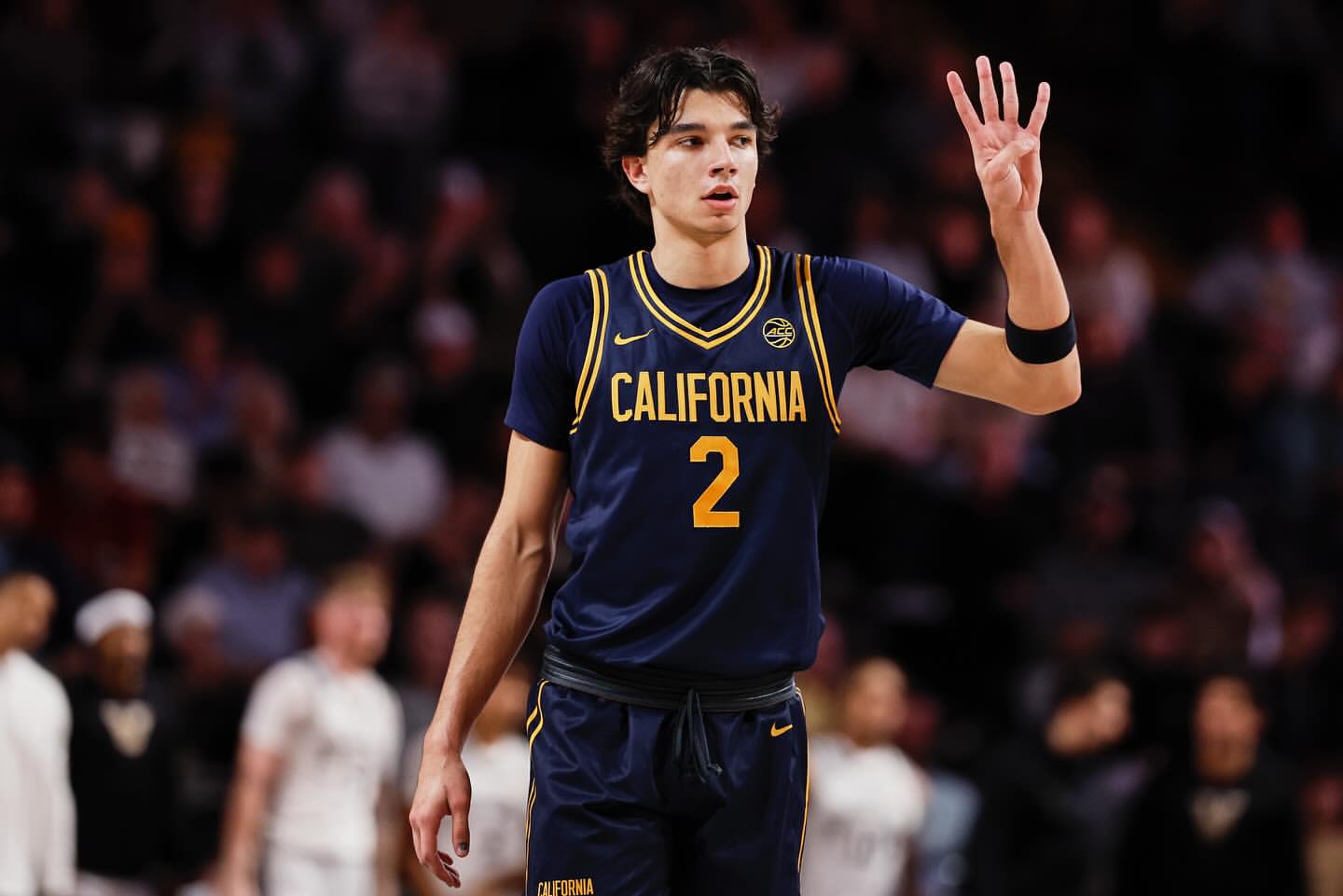 Male basketball player in blue and gold uniform holds up four fingers on court. Spectators blurred in background. Sports action.