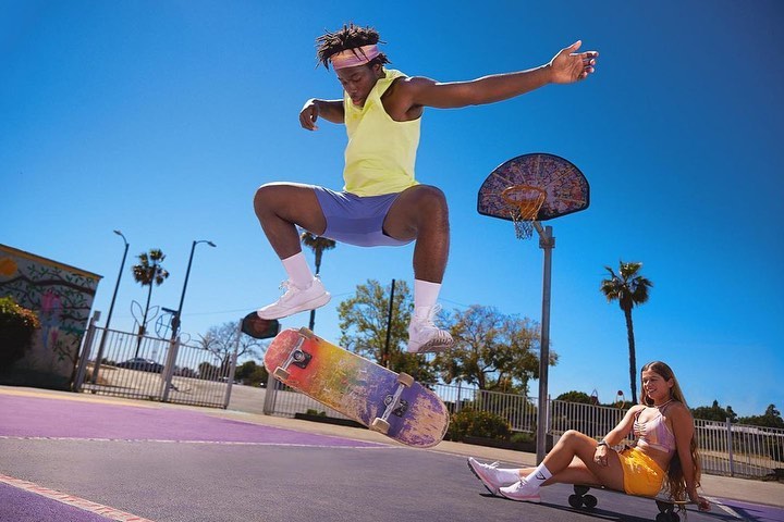 Athletic black man mid-air skateboarding in an urban park with a woman lounging on a skateboard under clear blue sky.
