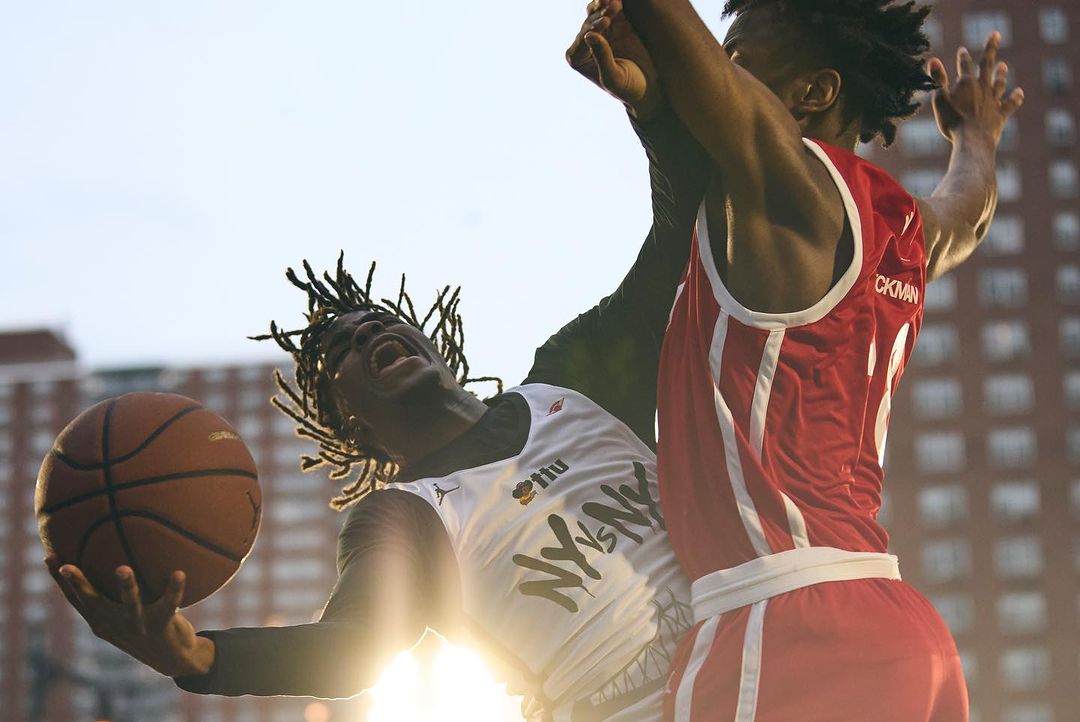 Two basketball players compete for the ball in a low-angle shot with a building in the background and sun flare, during daytime outdoors.