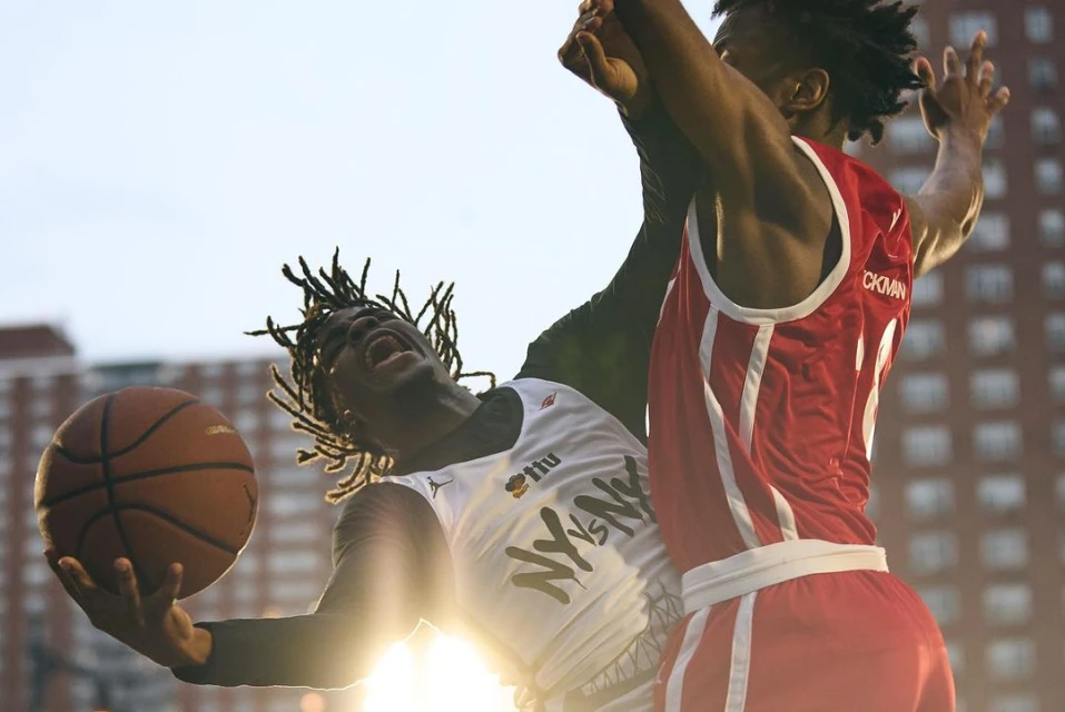 Two basketball players compete for the ball in a low-angle shot with a building in the background and sun flare, during daytime outdoors.