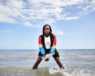 Tennis player Coco Gauff standing in shallow water wearing a colour-blocked athletic jacket and holding a tennis racket