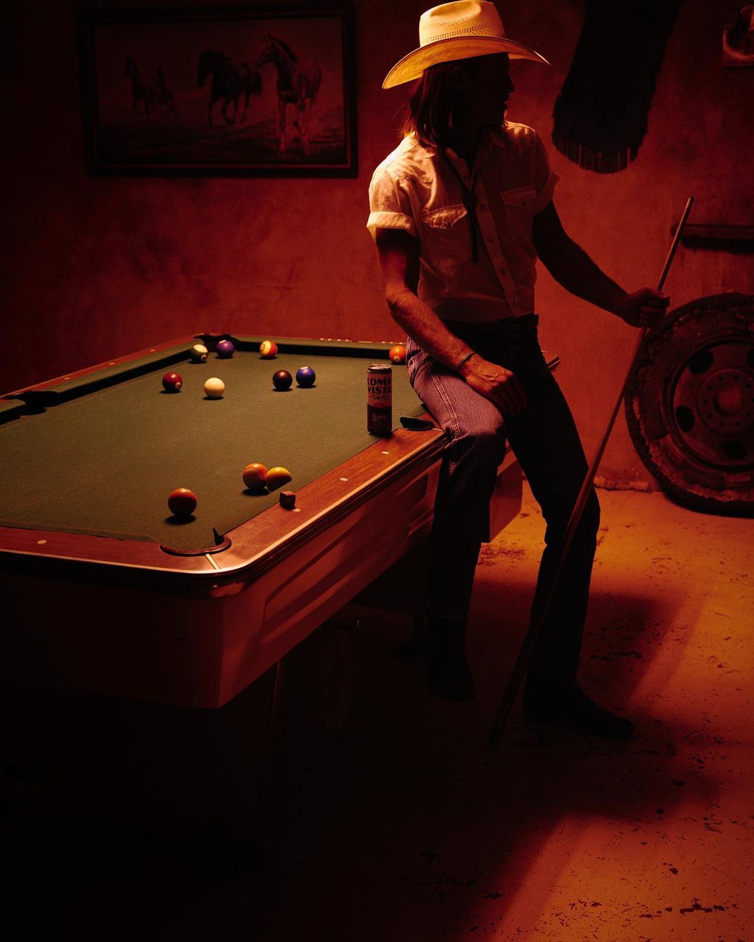 Man in cowboy hat sits on a billiards table, holding a pool cue, in a dimly lit room with vintage western decor and warm color grading.