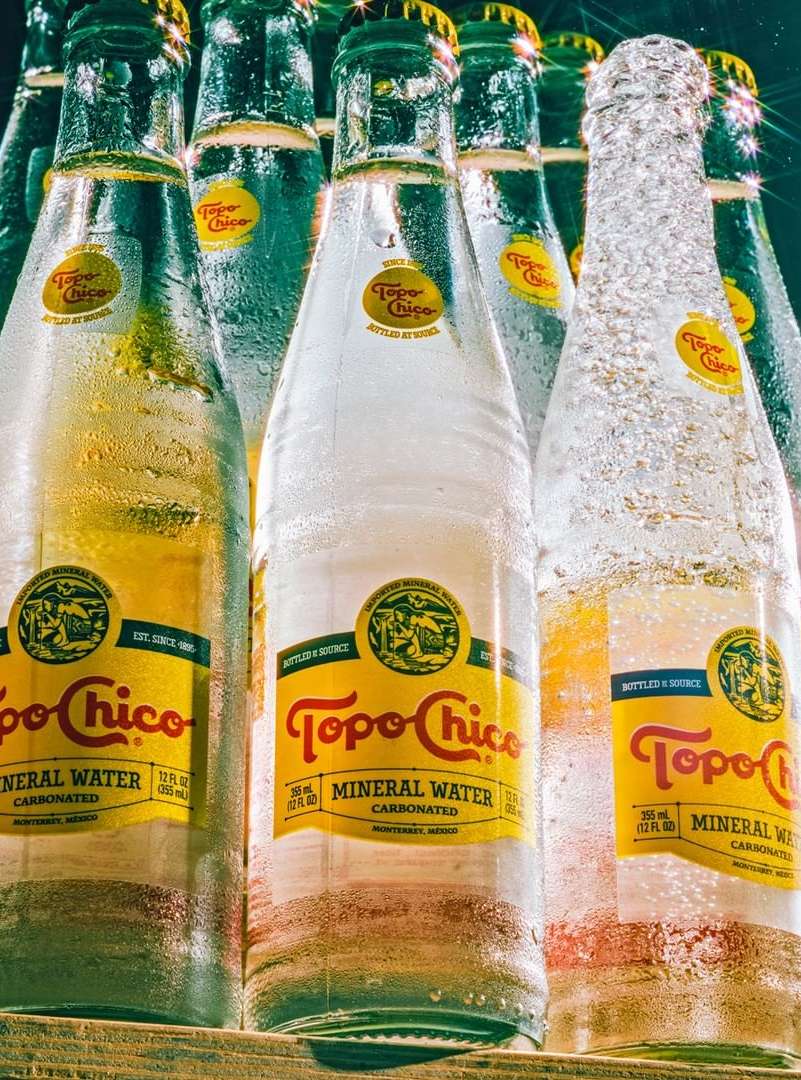 Multiple clear glass bottles of Topo Chico mineral water are densely arranged with visible condensation and distinctive yellow labels.