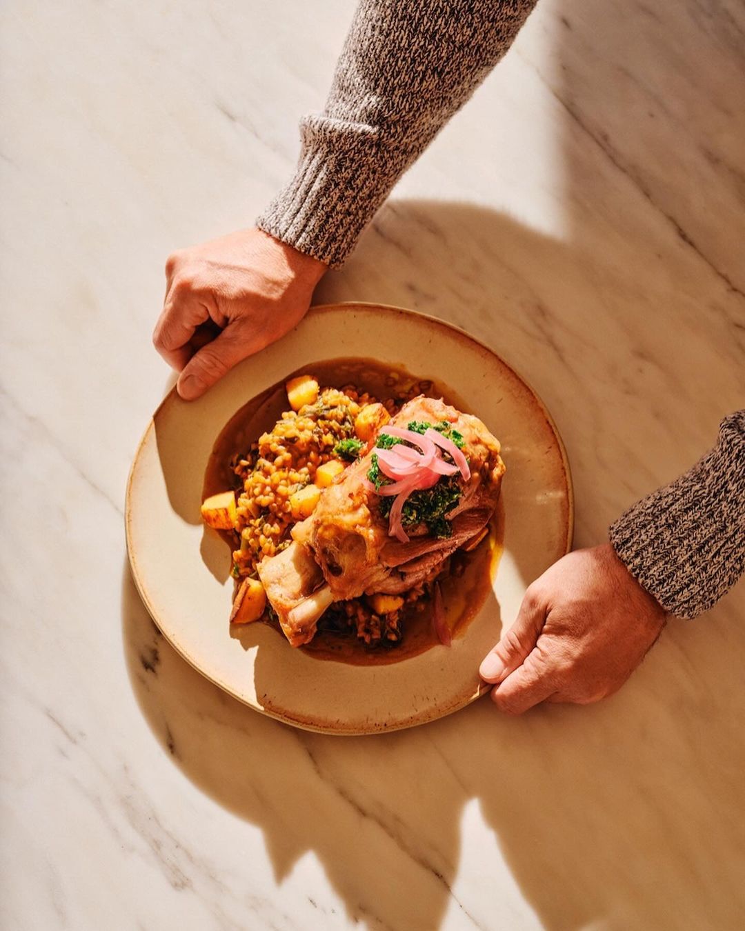 Overhead shot of a chef placing a plated lamb shank dish with lentils and pickled onions on a light marble surface.