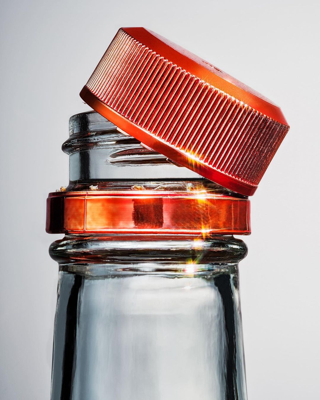 Close-up studio shot of a transparent glass bottle with a red, ridged cap partially covering the opening, set against a soft gradient background.