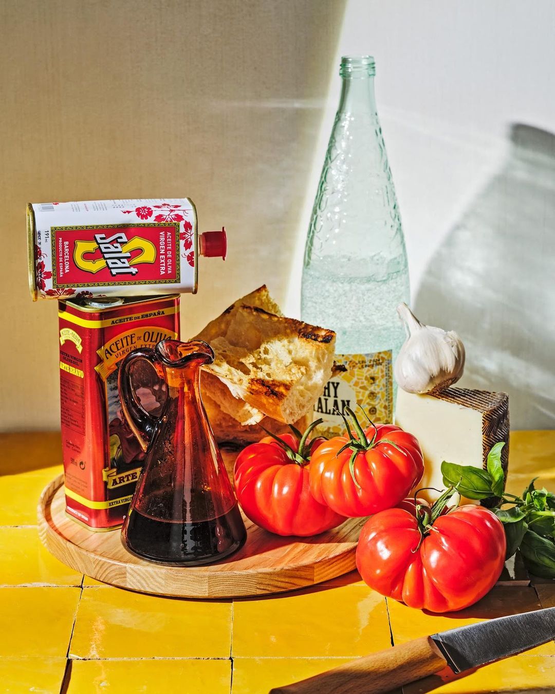 Still life featuring tomatoes, olive oil, bread, cheese, garlic, balsamic vinegar, and basil arranged on a yellow tiled surface.