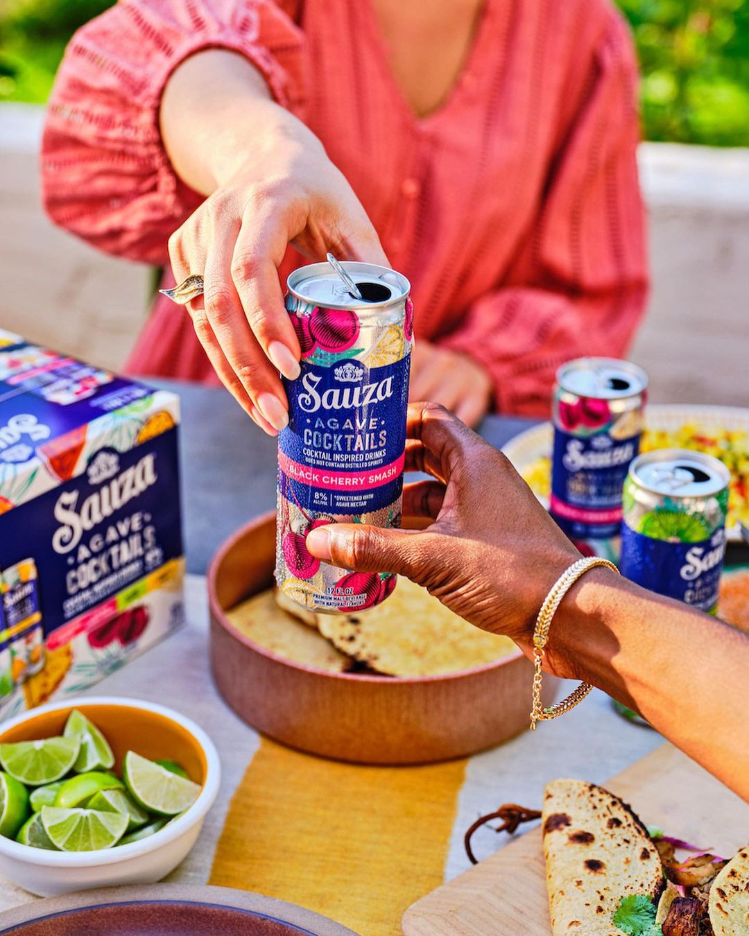 Two people exchanging a can of Sauza Agave Cocktail. Lime wedges, tortillas, and other cans are visible on a table in the background.