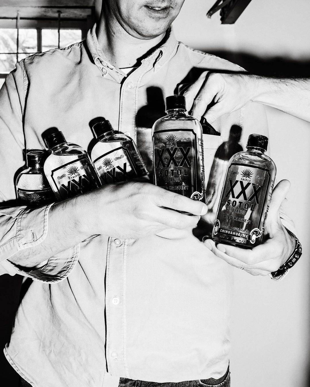 Person holding five bottles of XXX Sotol liquor indoors. The black and white portrait showcases the man's hands and the bottles against a window.