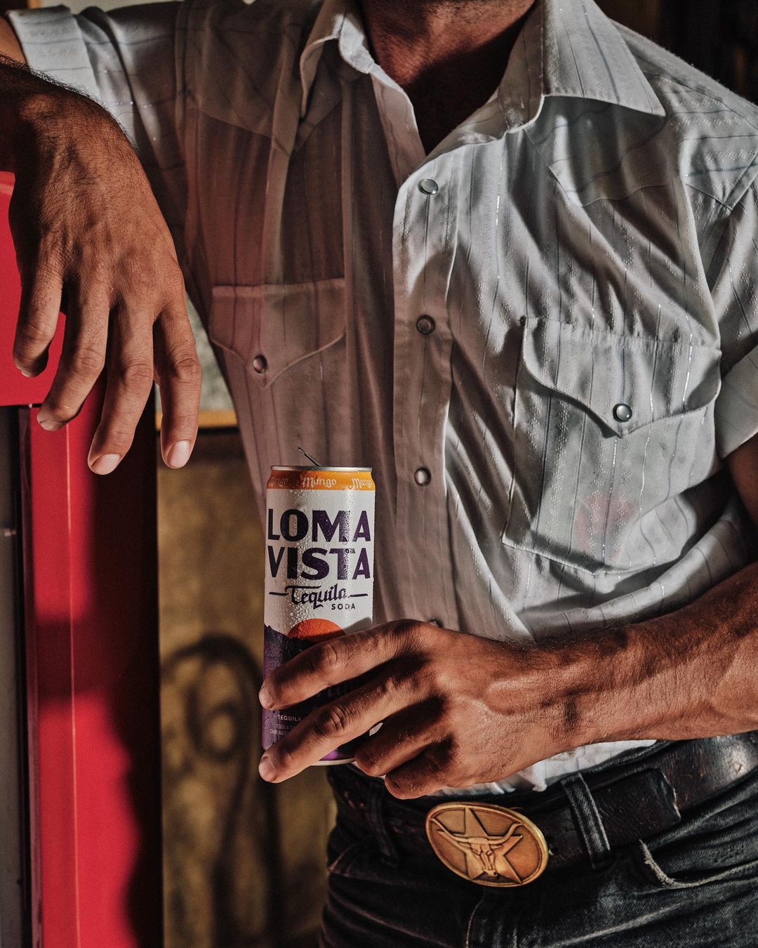Adult male in a white button-down shirt holding a can of Loma Vista Tequila Seltzer while leaning against a red wall indoors.