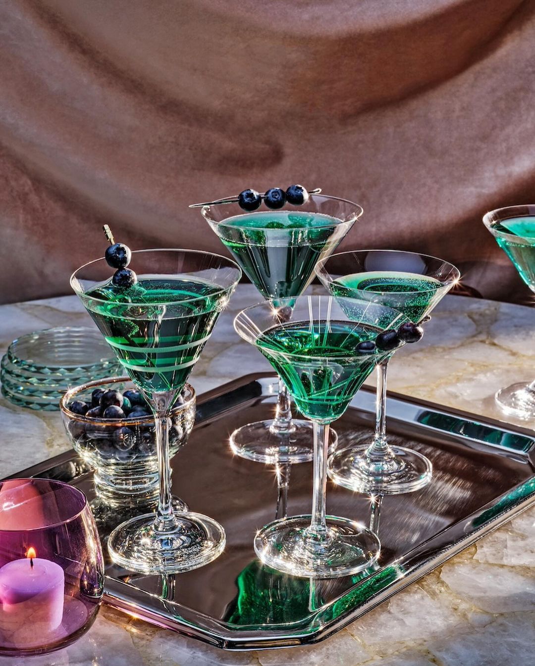 Five martini glasses filled with green cocktails and garnished with blueberries on a silver tray, warm lighting, and a gold backdrop.