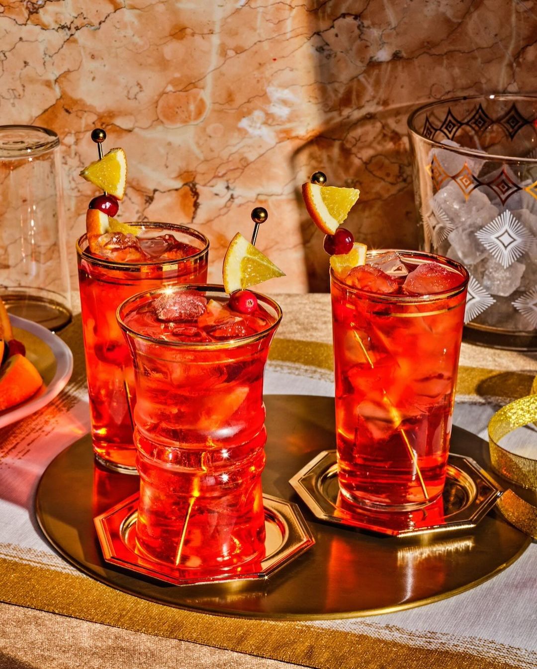 Three red cocktails with orange slices and cherries on a gold tray, illuminated by soft lighting against a marble backdrop.