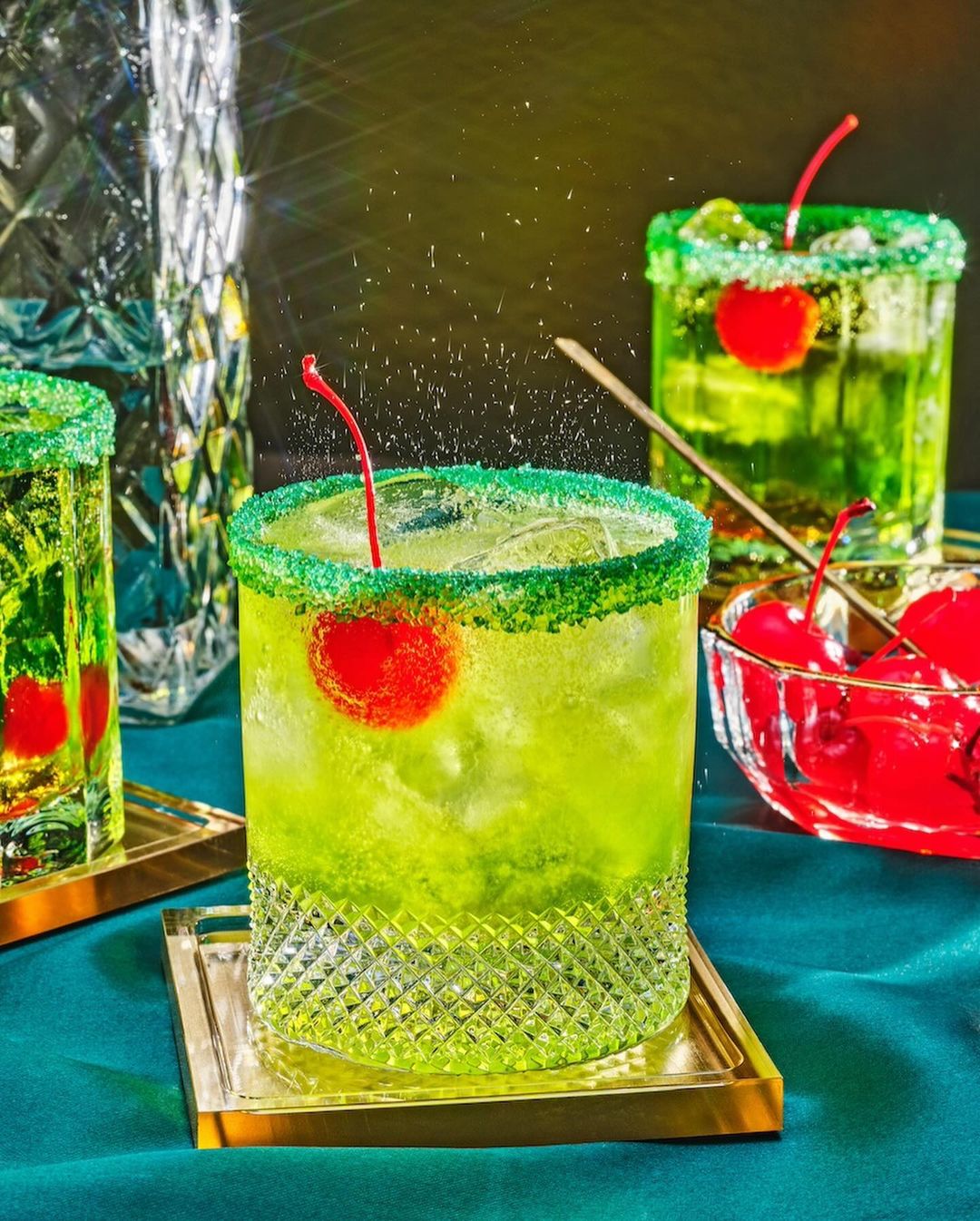 Three green cocktails are topped with cherries, served in decorative glasses with sugared rims, and set on gold coasters.