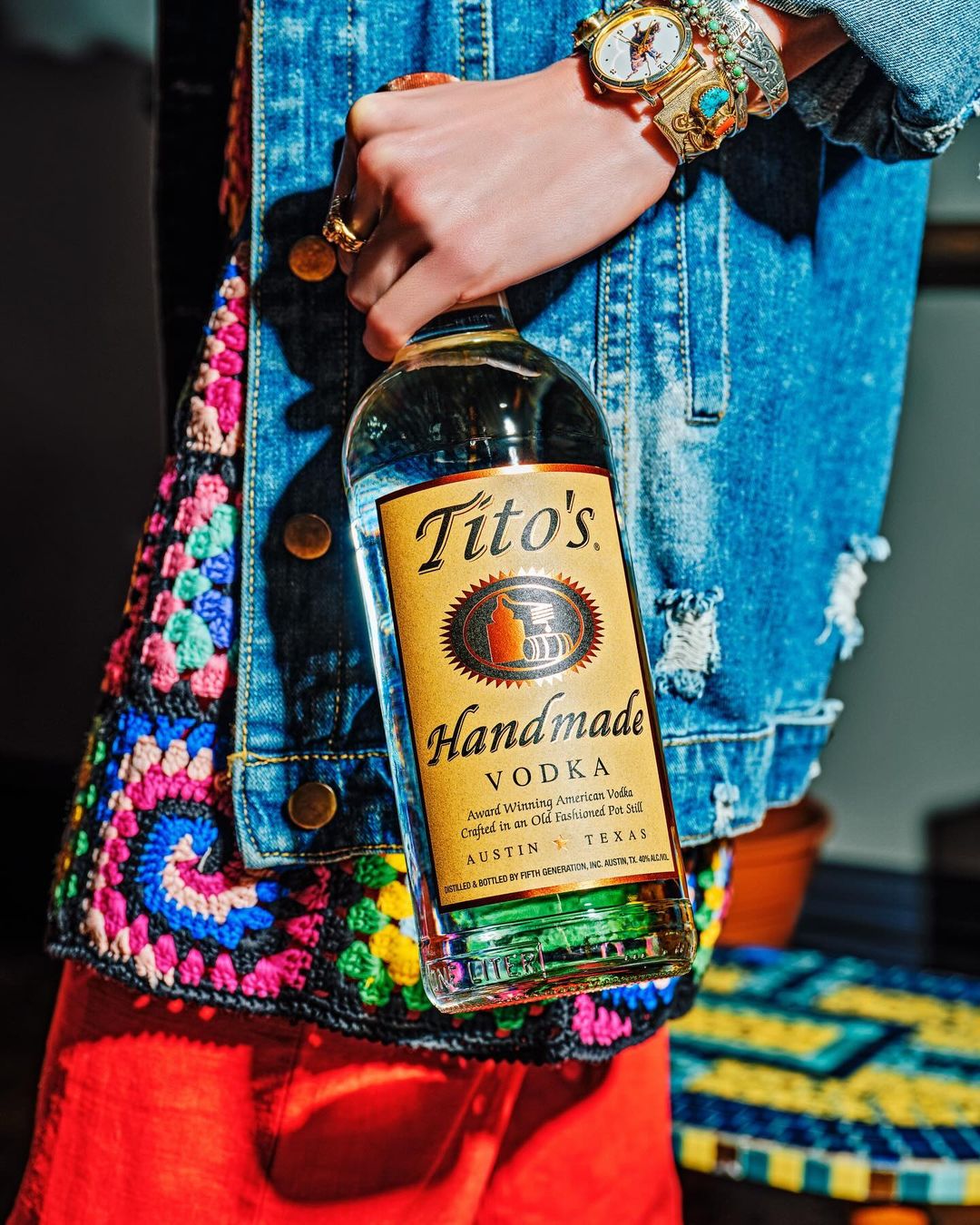 Close-up of a hand holding a bottle of Tito's Handmade Vodka, with a denim jacket, colorful crochet detail, and jewelry visible.