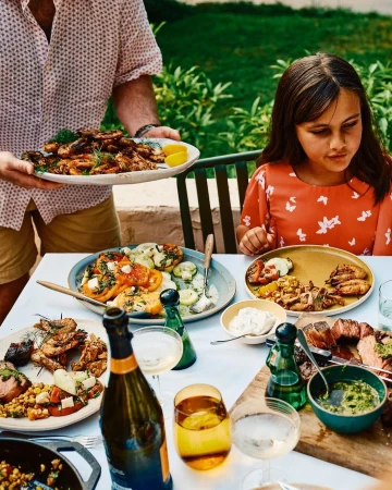 Child sits at a table laden with grilled food and drinks, served by an adult at a backyard gathering, with green foliage.