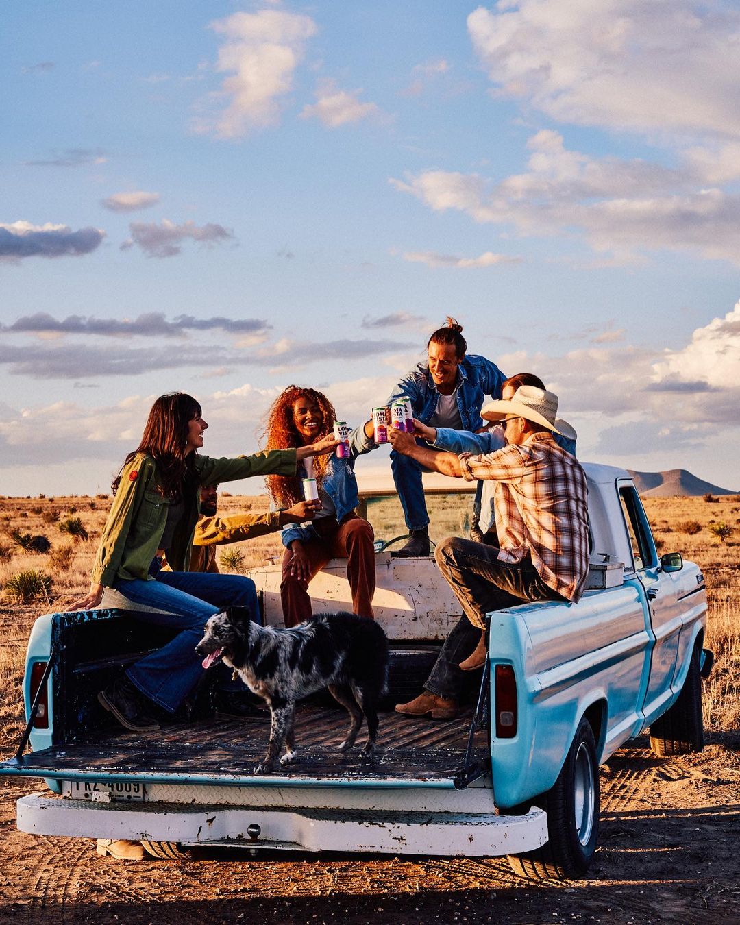 Four friends and a dog gather on the tailgate of a vintage pickup truck in a rural desert setting, toasting with beverage cans during sunset.