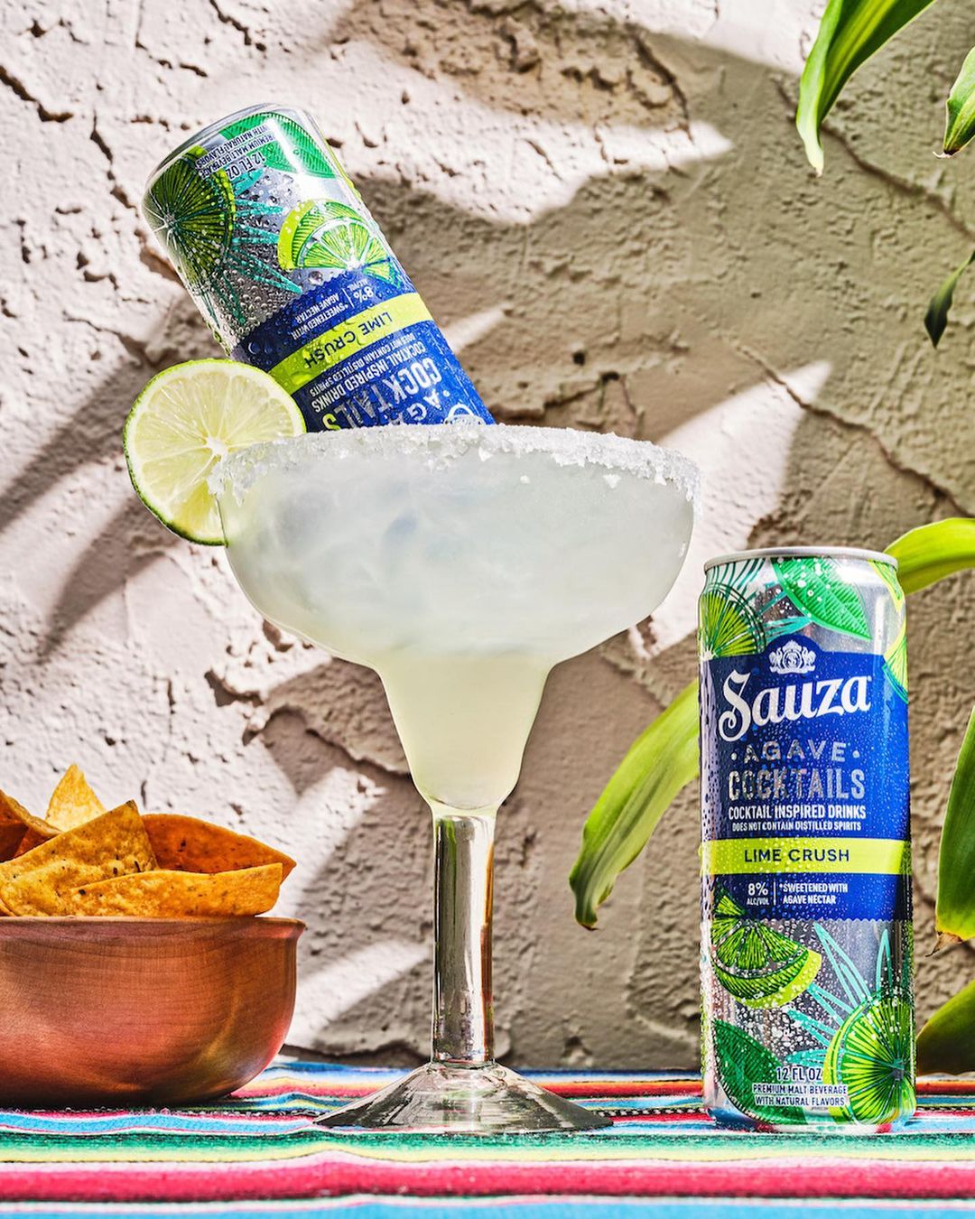 Margarita cocktail with lime slice, can of Sauza Lime Crush agave cocktail, and tortilla chips against textured wall and plants.