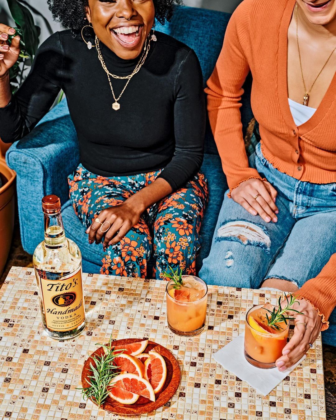 Two cheerful women lounge on a blue couch, enjoying orange cocktails on a mosaic table with vodka bottle, grapefruit, and rosemary.